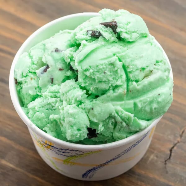 A white cup filled with scoops of bright green mint chocolate chip ice cream on a wooden table.