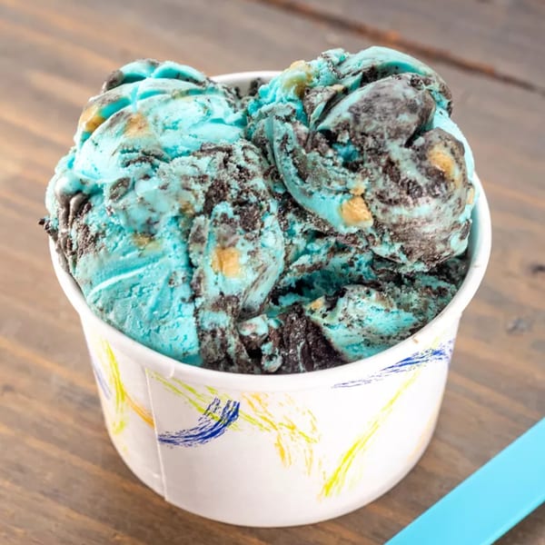 Scoops of blue ice cream with dark cookie crumbles and yellow chunks in a white cup.